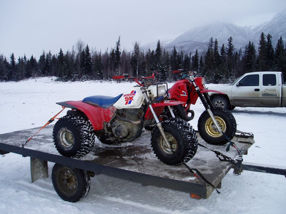 Two Honda SX three-wheelers on a trailer in the snow, with a pickup truck and mountains in the background