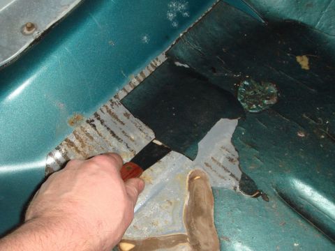 1996 Ford Bronco - A hand scrapes away rust and undercoating from a teal vehicle's interior floor.