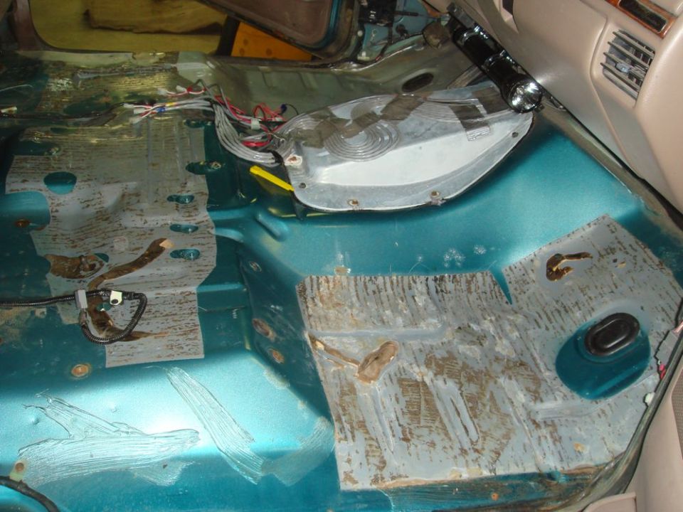 Interior floor of a vehicle with rust and exposed metal panels. Wiring is visible