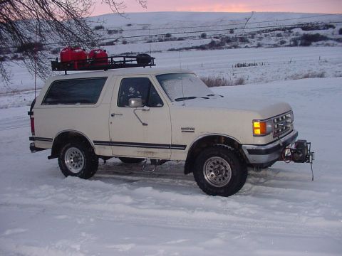 White 1987 Ford Bronco with a roof rack carrying red gas cans, parked in snow