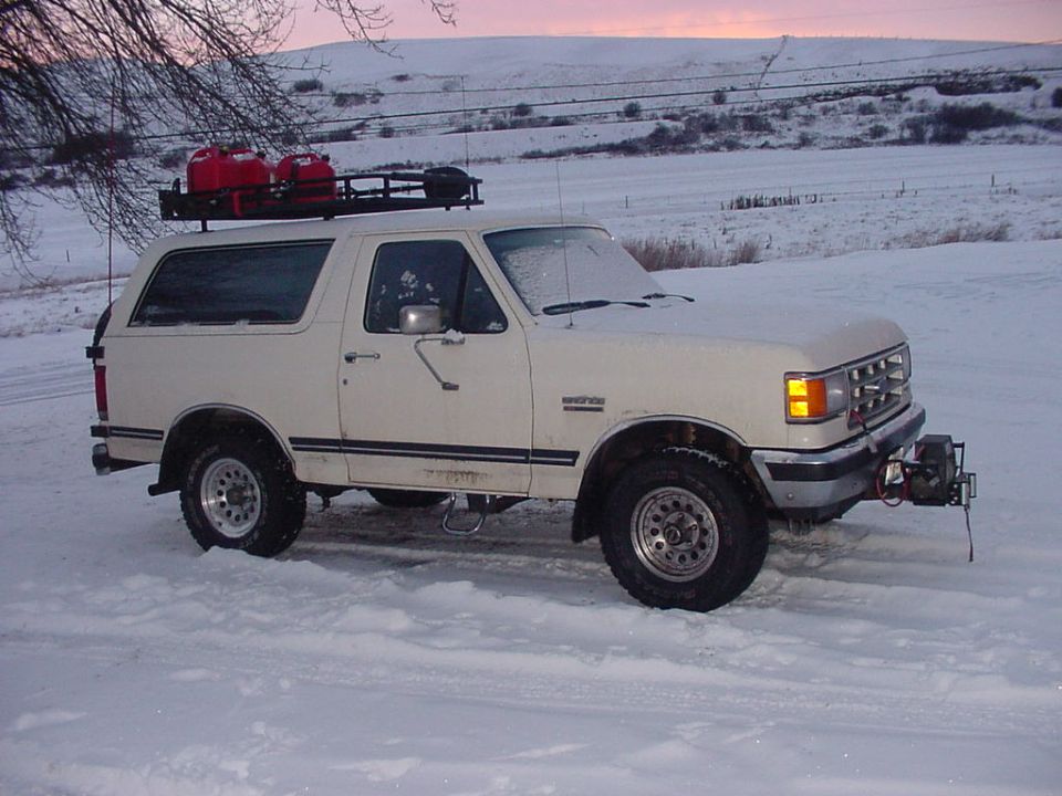 White 1987 Ford Bronco with a roof rack carrying red gas cans, parked in snow