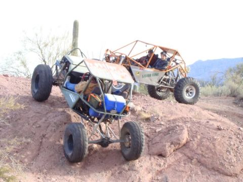 1987 Suzuki Samurai - Two custom off-road vehicles navigate a steep, rocky desert terrain under a bright sky.