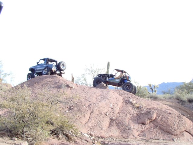 Blue 1987 Suzuki Samurai with a spare tire on the back, climbing a dirt hill. Another off-road buggy is to its right