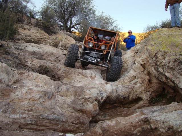 Orange off-road buggy with large tires navigating a rocky incline. Two people are visible inside the buggy