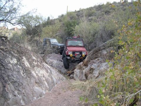 1987 Suzuki Samurai - Two off-road vehicles tackle a challenging, rocky path in a desert landscape.