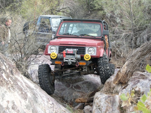 Red 1987 Suzuki Samurai off-roading over rocks, with a black SUV behind it