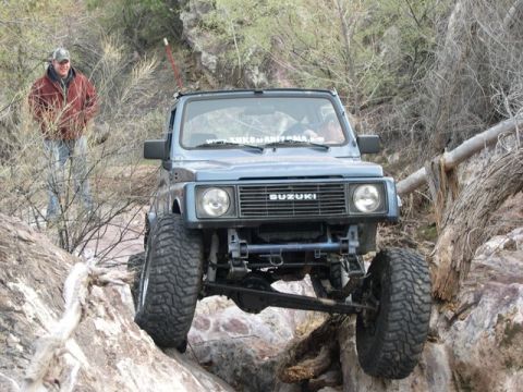 1987 Suzuki Samurai - A blue 1987 Suzuki Samurai is shown off-roading over a rocky landscape, with a man standing nearby.