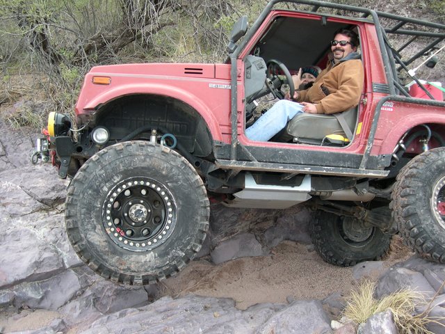 Red 1987 Suzuki Samurai with large tires on rocky terrain, driver giving thumbs up