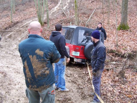 2002 Ford Sport Trac - A red Jeep Wrangler is stuck in a muddy, wooded area with several people observing the situation.