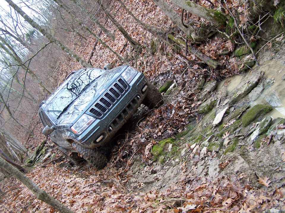 Jeep Grand Cherokee navigating a muddy, wooded trail with steep inclines and rocky terrain
