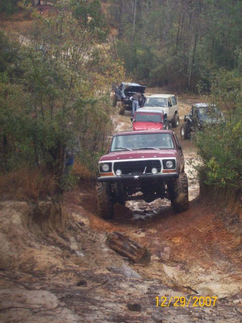 1978 Ford Bronco - Several off-road vehicles navigate a challenging, muddy track through a wooded area.