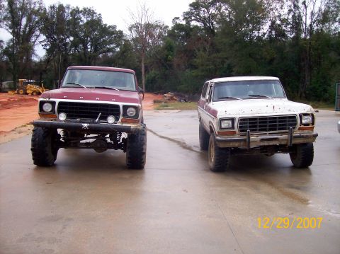 1978 Ford Bronco - Two lifted Ford trucks are parked on a wet surface, with trees and construction equipment in the background.