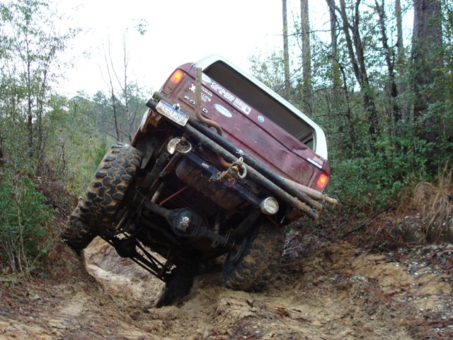 Maroon Ford Bronco with white top flexing at a steep angle on a muddy trail, rear underside view