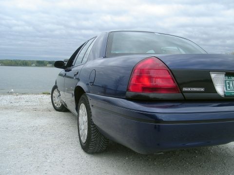 2003 Ford Crown Victoria - Rear view of a blue 2003 Ford Crown Victoria Police Interceptor parked by a body of water under a cloudy sky.