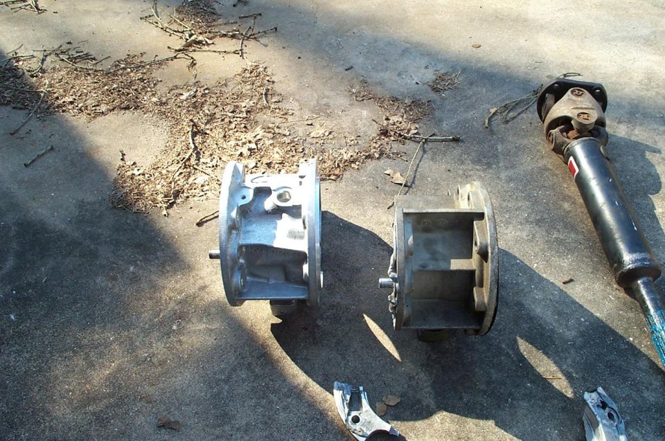 Two metal automotive housings and a driveshaft lie on a concrete surface with scattered leaves