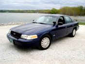 2003 Ford Crown Victoria - A blue 2003 Ford Crown Victoria is parked on a gravel lot next to a calm lake.