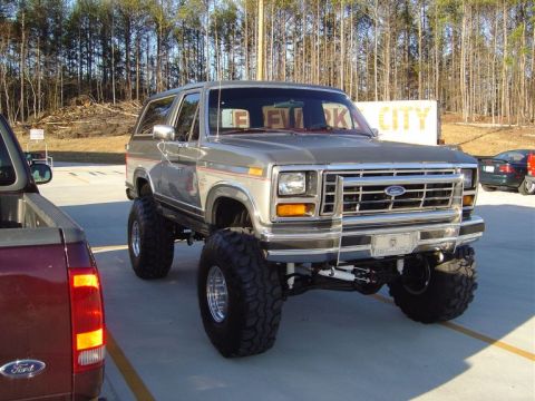 1986 Ford Bronco - A lifted silver Ford Bronco is parked next to a red pickup truck.