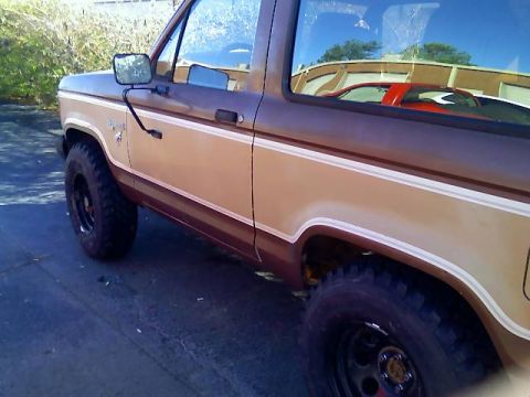 1984 Ford Bronco II - The side profile of a two-tone brown and tan 1984 Ford Bronco II is visible, with off-road tires and a reflection of another vehicle in the…
