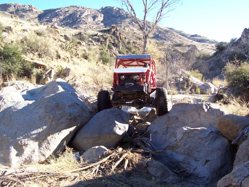 Red off-road vehicle with roll cage navigating large rocks on a sunny day