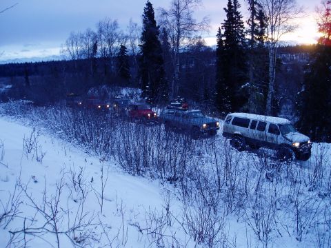 2000 Ford E350 - A line of off-road vehicles navigates a snow-covered path in a wooded area at dusk.