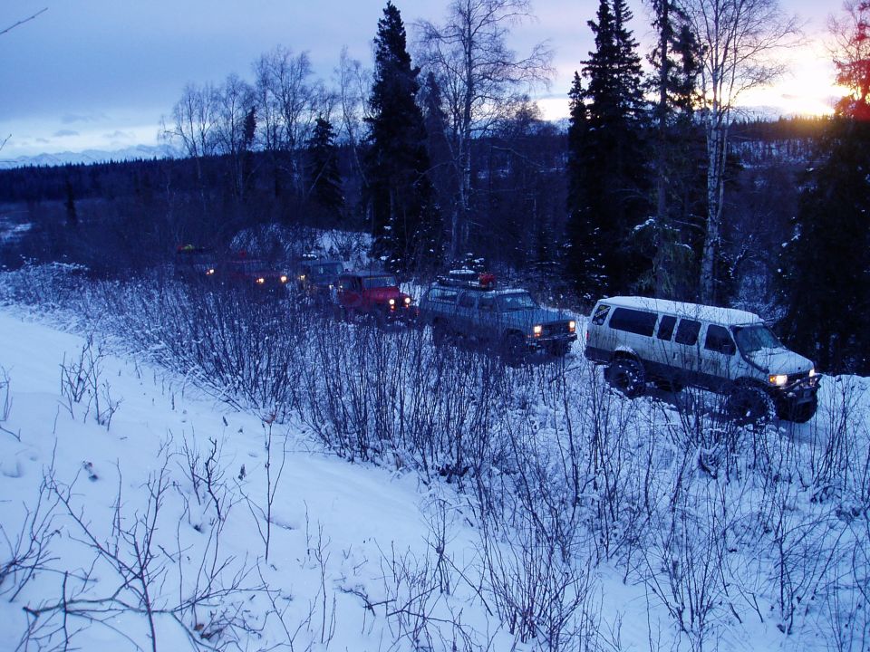 Several SUVs and vans driving in a line on a snowy trail through a forest