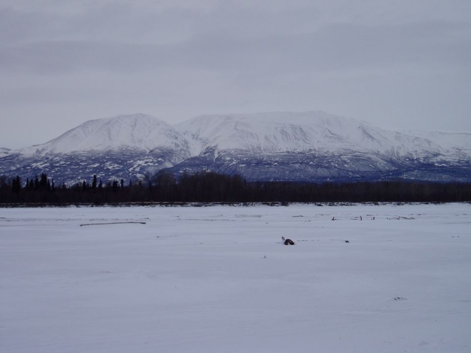 Snowy mountains and a vast, flat, snow-covered plain with a few scattered objects