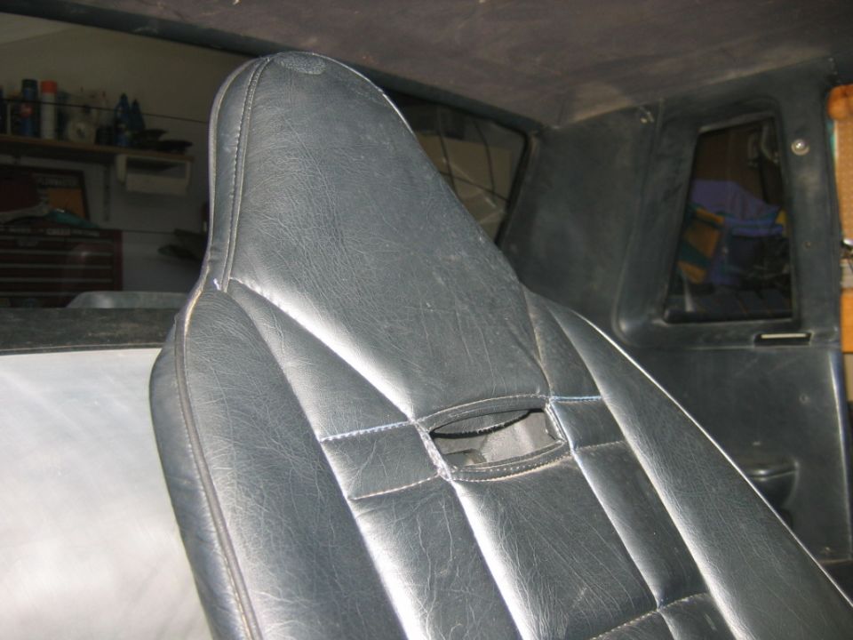 Close-up of a black vinyl car seat with stitching detail. The headrest is visible, and a small opening is in the seat back. Interior panels and a window are in the background
