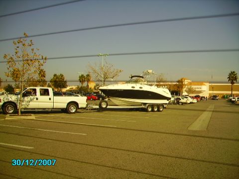 1990 Ford Bronco - A white pickup truck tows a boat on a trailer through a parking lot on a sunny day.
