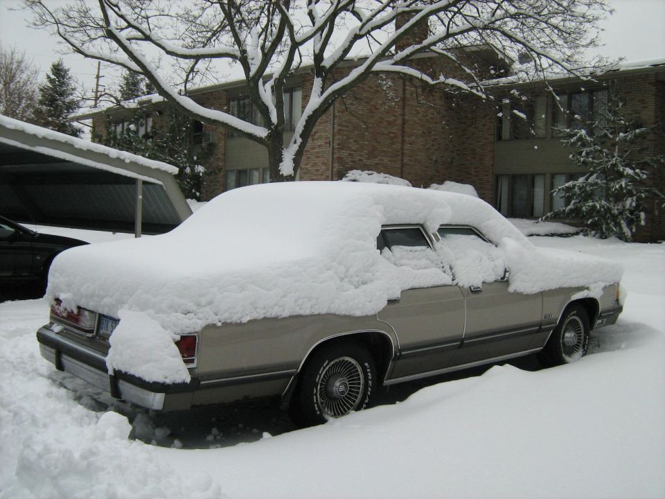 Snow-covered tan two-door car parked by a brick building
