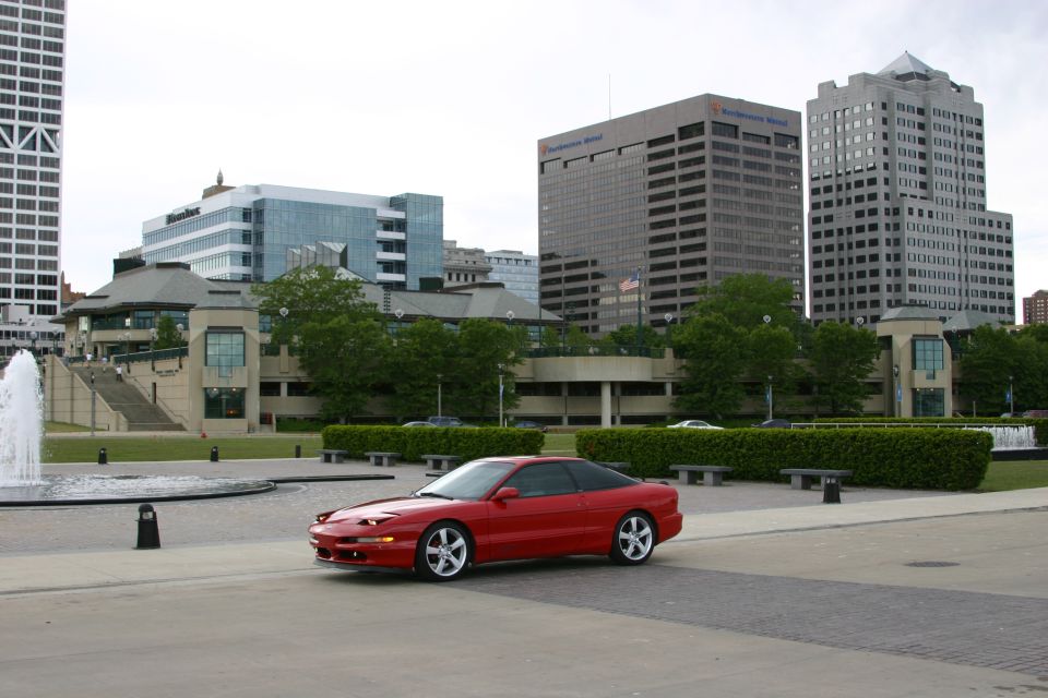 Red Ford Probe coupe parked before a city skyline and fountain