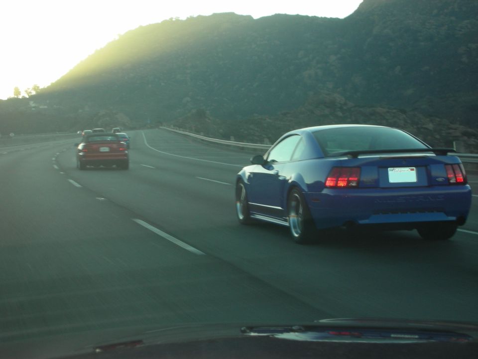 Blue Ford Mustang driving on highway, sun setting behind mountains