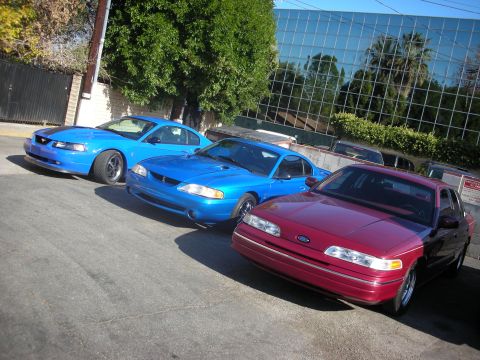 1992 Ford Crown Victoria - Three blue Ford Mustangs and a red Ford Crown Victoria are parked in a lot.