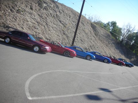 1992 Ford Crown Victoria - A line of cars, including a maroon 1992 Ford Crown Victoria, are parked on a sloped asphalt surface next to a dirt embankment.