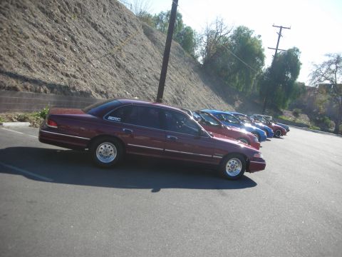 1992 Ford Crown Victoria - A line of sedans, including a maroon 1992 Ford Crown Victoria, parked on asphalt with a dirt embankment in the background.