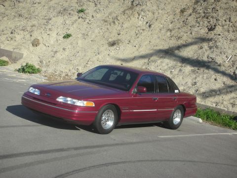 1992 Ford Crown Victoria - A maroon 1992 Ford Crown Victoria is parked on a street with a dirt embankment in the background.