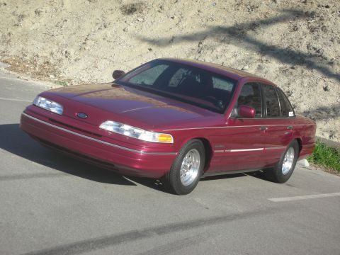 1992 Ford Crown Victoria - A maroon 1992 Ford Crown Victoria sedan is parked on an asphalt surface with a dirt embankment in the background.