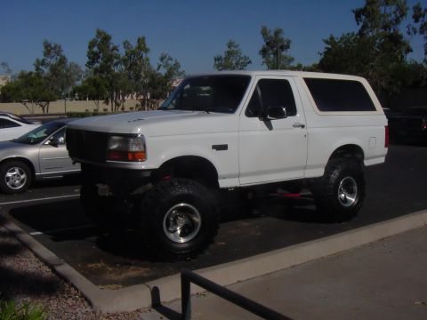1992 Ford Bronco - A lifted 1992 Ford Bronco is parked in a lot next to a silver sedan under a clear sky.