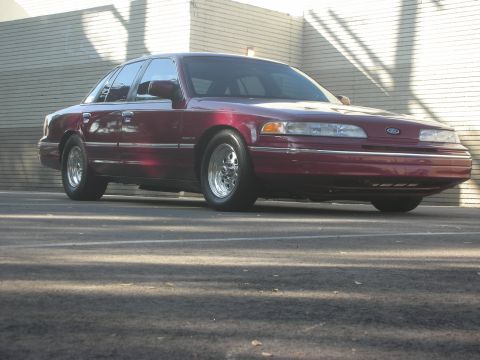 1992 Ford Crown Victoria - A maroon 1992 Ford Crown Victoria is parked in front of a brick wall, casting long shadows.