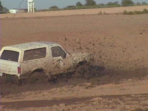 1992 Ford Bronco - A white SUV is stuck in a mud pit, with mud spraying from its wheels.