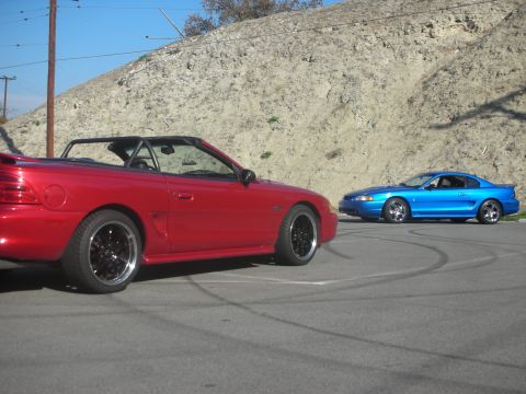 1992 Ford Crown Victoria - Two sporty coupes, a red convertible and a blue coupe, are parked on asphalt with tire marks visible.