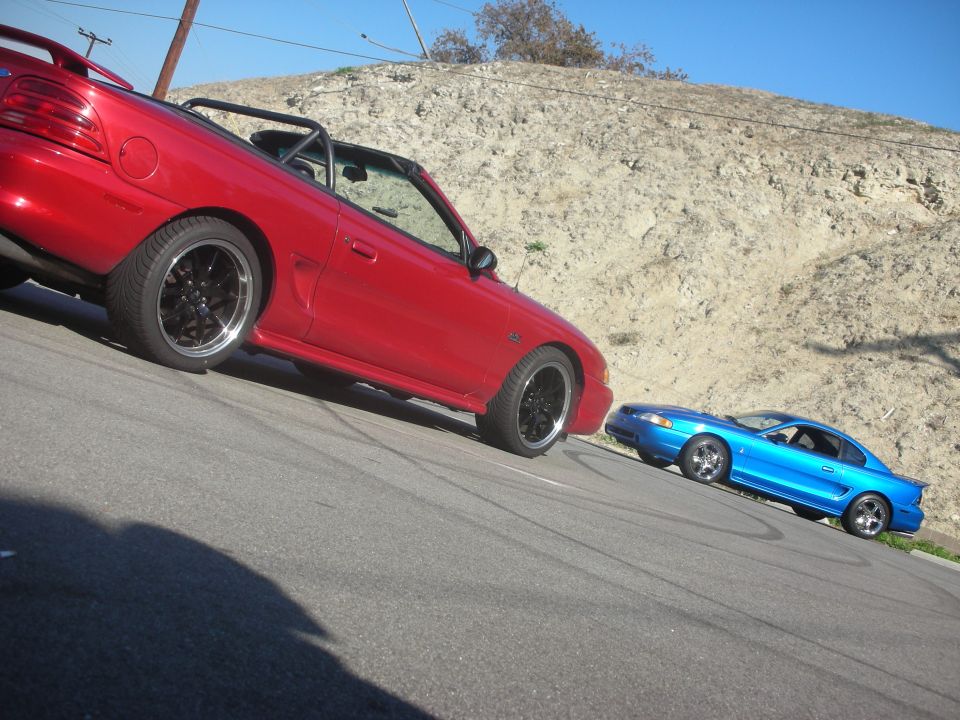 Red convertible Ford Mustang and blue coupe Ford Mustang parked on a street with a hill in the background
