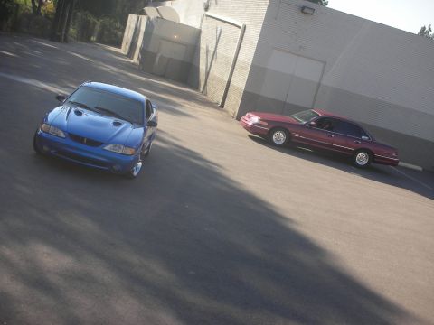 1992 Ford Crown Victoria - Two cars, a blue Ford Mustang and a maroon Ford Crown Victoria, are parked in an asphalt lot next to a building.