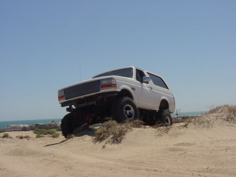 1992 Ford Bronco - A white 1992 Ford Bronco navigates sandy terrain under a clear blue sky.