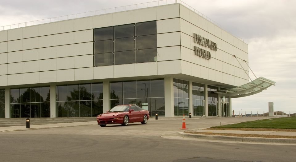Red 1993 Ford Probe driving in front of a modern white building with "DISCOVER WORLD" signage