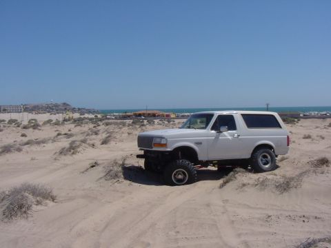 1992 Ford Bronco - A white Ford Bronco is parked on a sandy terrain with the ocean in the background.