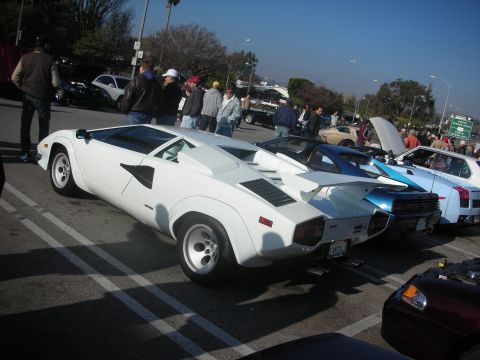 1998 Ford Crown Victoria - A white Lamborghini Countach is parked in a lot with several other vehicles and people milling around.