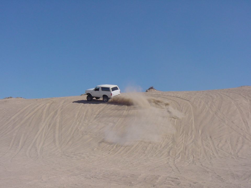 White 1992 Ford Bronco kicking up dust while driving on a sand dune under a clear blue sky