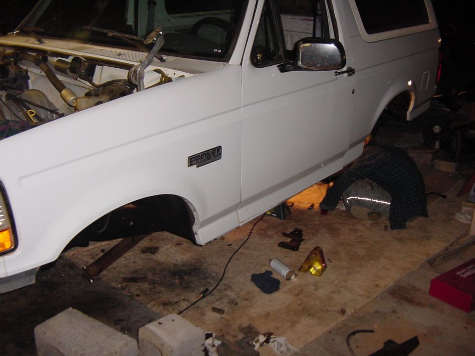 White 1992 Ford Bronco on jack stands, front fender and engine bay visible. Someone is working underneath