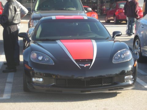1998 Ford Crown Victoria - A black Chevrolet Corvette with distinctive red and silver racing stripes is parked in a lot, with other vehicles and people nearby.