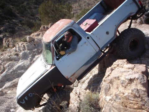1989 Ford Bronco - A white pickup truck is shown off-roading on a steep, rocky incline, with the driver visible inside.